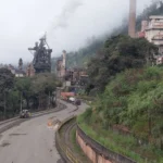 Imagem panorâmica da ArcelorMittal Monlevade, em João Monlevade (MG), mostrando as instalações da usina siderúrgica, chaminés, estruturas industriais, via de acesso e linha férrea em primeiro plano, com áreas de vegetação ao redor.