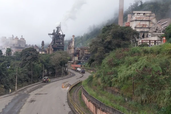 Imagem panorâmica da ArcelorMittal Monlevade, em João Monlevade (MG), mostrando as instalações da usina siderúrgica, chaminés, estruturas industriais, via de acesso e linha férrea em primeiro plano, com áreas de vegetação ao redor.