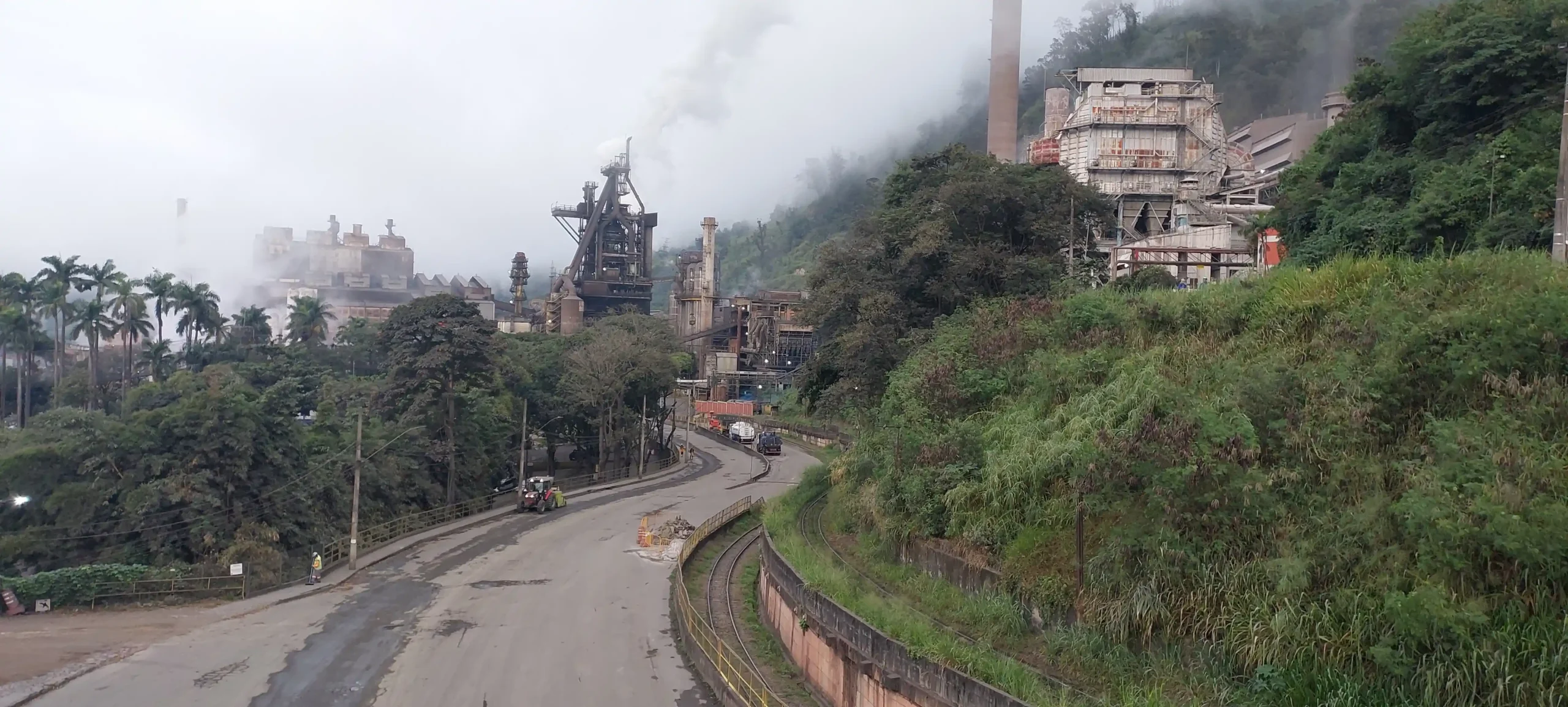 Imagem panorâmica da ArcelorMittal Monlevade, em João Monlevade (MG), mostrando as instalações da usina siderúrgica, chaminés, estruturas industriais, via de acesso e linha férrea em primeiro plano, com áreas de vegetação ao redor.