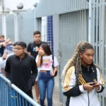 Grupo de jovens estudantes segue por um corredor externo de universidade ou escola. Em destaque, uma estudante segura um celular enquanto caminha. A imagem representa a oportunidade de ingresso em uma Graduação gratuita de Direito que será ofertada em João Monlevade.