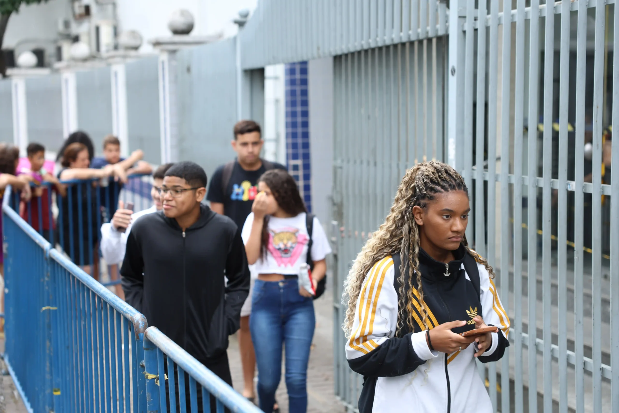 Grupo de jovens estudantes segue por um corredor externo de universidade ou escola. Em destaque, uma estudante segura um celular enquanto caminha. A imagem representa a oportunidade de ingresso em uma Graduação gratuita de Direito que será ofertada em João Monlevade.