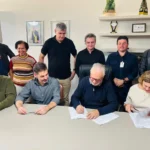 Grupo de autoridades reunido em uma sala durante a assinatura de documentos relacionados à construção da nova quadra da Escola Municipal Cônego José Higino de Freitas. O prefeito assina o contrato enquanto gestores, vereadores e representantes da educação acompanham a cerimônia.