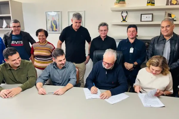 Grupo de autoridades reunido em uma sala durante a assinatura de documentos relacionados à construção da nova quadra da Escola Municipal Cônego José Higino de Freitas. O prefeito assina o contrato enquanto gestores, vereadores e representantes da educação acompanham a cerimônia.