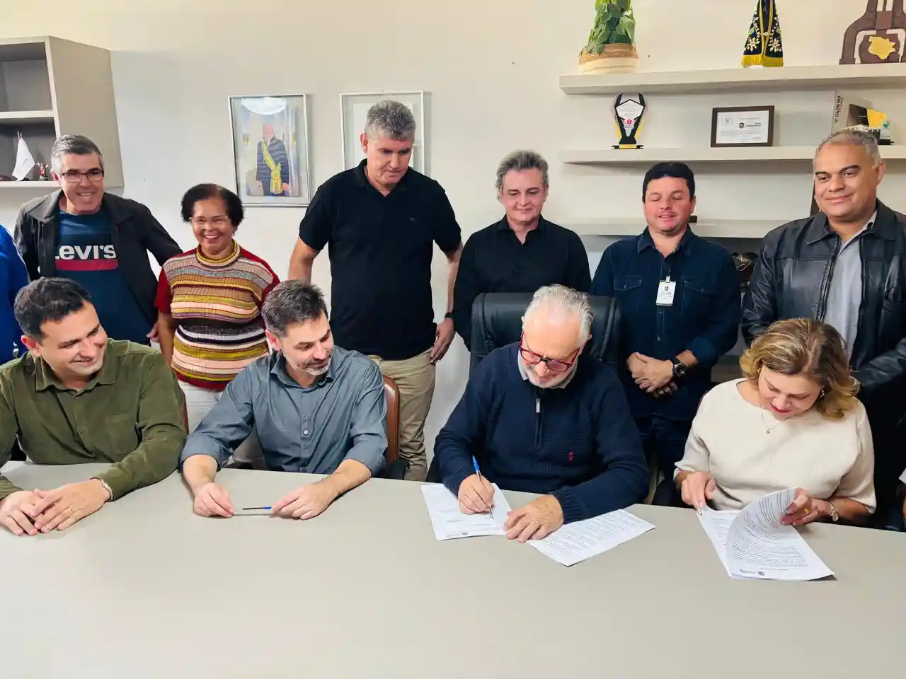 Grupo de autoridades reunido em uma sala durante a assinatura de documentos relacionados à construção da nova quadra da Escola Municipal Cônego José Higino de Freitas. O prefeito assina o contrato enquanto gestores, vereadores e representantes da educação acompanham a cerimônia.