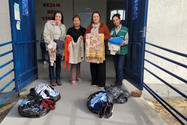 Quatro mulheres posam na entrada de um prédio enquanto exibem agasalhos, cobertores e roupas doadas. No chão, há sacolas com mais peças de vestuário e calçados. A ação faz parte da Campanha do Agasalho 2026, destinada ao atendimento de pessoas em situação de vulnerabilidade social em João Monlevade.