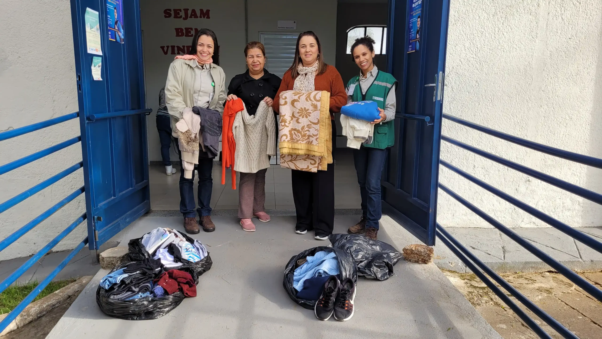 Quatro mulheres posam na entrada de um prédio enquanto exibem agasalhos, cobertores e roupas doadas. No chão, há sacolas com mais peças de vestuário e calçados. A ação faz parte da Campanha do Agasalho 2026, destinada ao atendimento de pessoas em situação de vulnerabilidade social em João Monlevade.