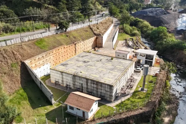 Imagem aérea da obra de saneamento ETE Carneirinhos, em João Monlevade, mostrando os tanques da estação de tratamento de esgoto, prédios de operação, áreas gramadas e um córrego ao lado da estrutura. A unidade foi construída para o tratamento de esgoto e fortalecer o saneamento básico na cidade.