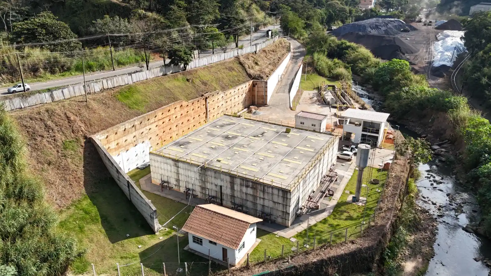 Imagem aérea da obra de saneamento ETE Carneirinhos, em João Monlevade, mostrando os tanques da estação de tratamento de esgoto, prédios de operação, áreas gramadas e um córrego ao lado da estrutura. A unidade foi construída para o tratamento de esgoto e fortalecer o saneamento básico na cidade.