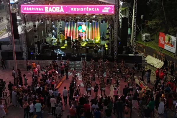 Vista aérea noturna da Praça do Povo durante o Festival Baobá, de cultura afro-brasileira. Um grande palco iluminado com luzes coloridas recebe apresentações culturais, enquanto dezenas de pessoas acompanham o evento.
