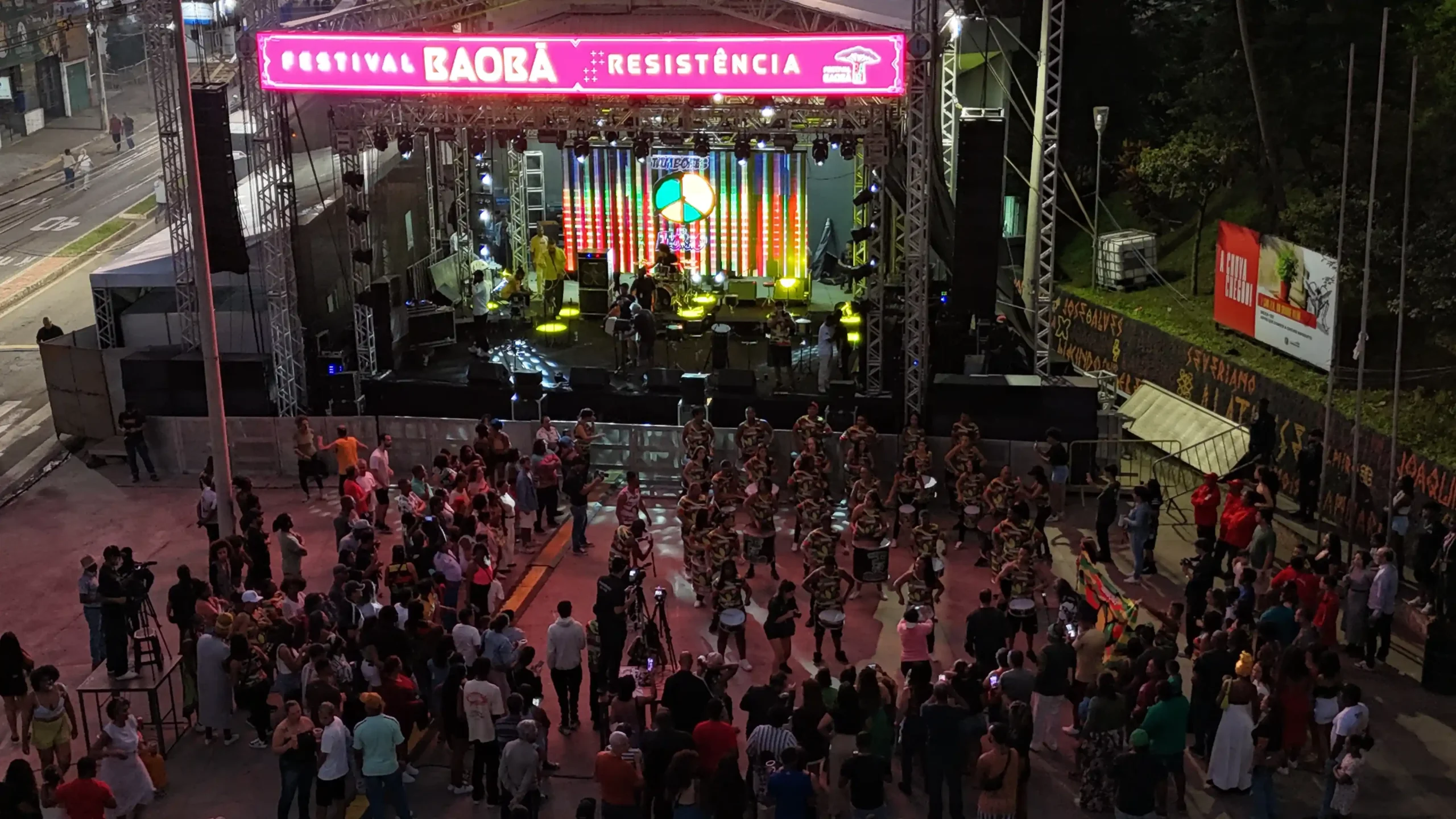 Vista aérea noturna da Praça do Povo durante o Festival Baobá, de cultura afro-brasileira. Um grande palco iluminado com luzes coloridas recebe apresentações culturais, enquanto dezenas de pessoas acompanham o evento.