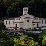 Patrimônio histórico de João Monlevade, a Igreja São José Operário aparece cercada por mata nativa, com fachada clara, torre central e telhados avermelhados, em uma paisagem que evidencia sua relevância cultural e arquitetônica.