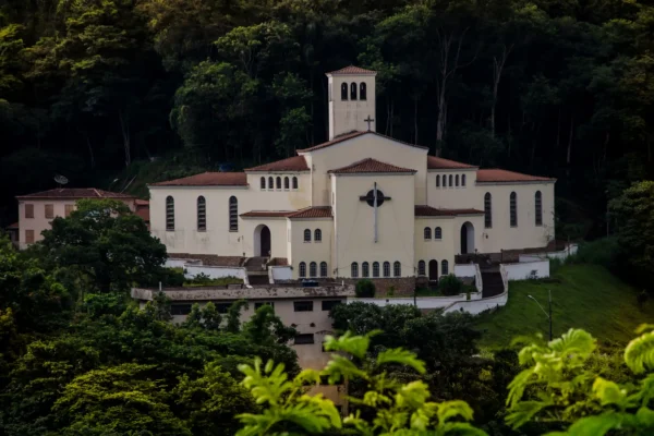 Patrimônio histórico de João Monlevade, a Igreja São José Operário aparece cercada por mata nativa, com fachada clara, torre central e telhados avermelhados, em uma paisagem que evidencia sua relevância cultural e arquitetônica.