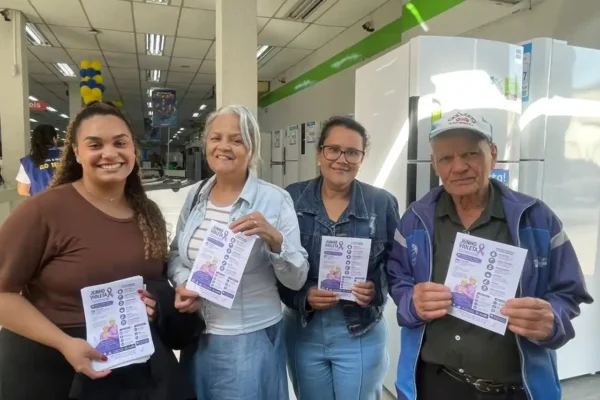 Quatro pessoas seguram panfletos da campanha Junho Violeta dentro de uma loja de eletrodomésticos no bairro Carneirinhos, em João Monlevade. A ação faz parte das atividades de conscientização sobre o combate à violência contra a pessoa idosa e a divulgação de seus direitos.