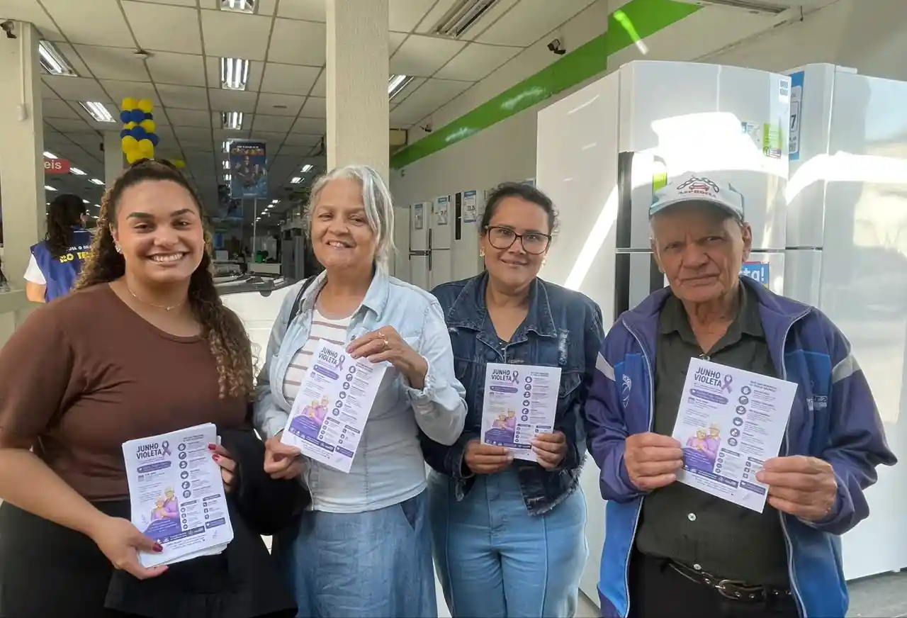Quatro pessoas seguram panfletos da campanha Junho Violeta dentro de uma loja de eletrodomésticos no bairro Carneirinhos, em João Monlevade. A ação faz parte das atividades de conscientização sobre o combate à violência contra a pessoa idosa e a divulgação de seus direitos.