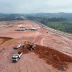 Vista aérea das obras do futuro Ponto de Parada e Descanso para caminhoneiros em Bom Jesus do Amparo, na BR-381, em Minas Gerais. Máquinas e caminhões realizam serviços de terraplanagem em uma ampla área de terra vermelha ao lado da rodovia, cercada por vegetação e colinas ao fundo.