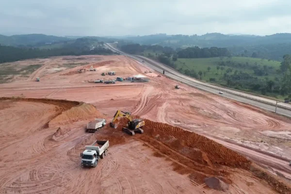 Vista aérea das obras do futuro Ponto de Parada e Descanso para caminhoneiros em Bom Jesus do Amparo, na BR-381, em Minas Gerais. Máquinas e caminhões realizam serviços de terraplanagem em uma ampla área de terra vermelha ao lado da rodovia, cercada por vegetação e colinas ao fundo.