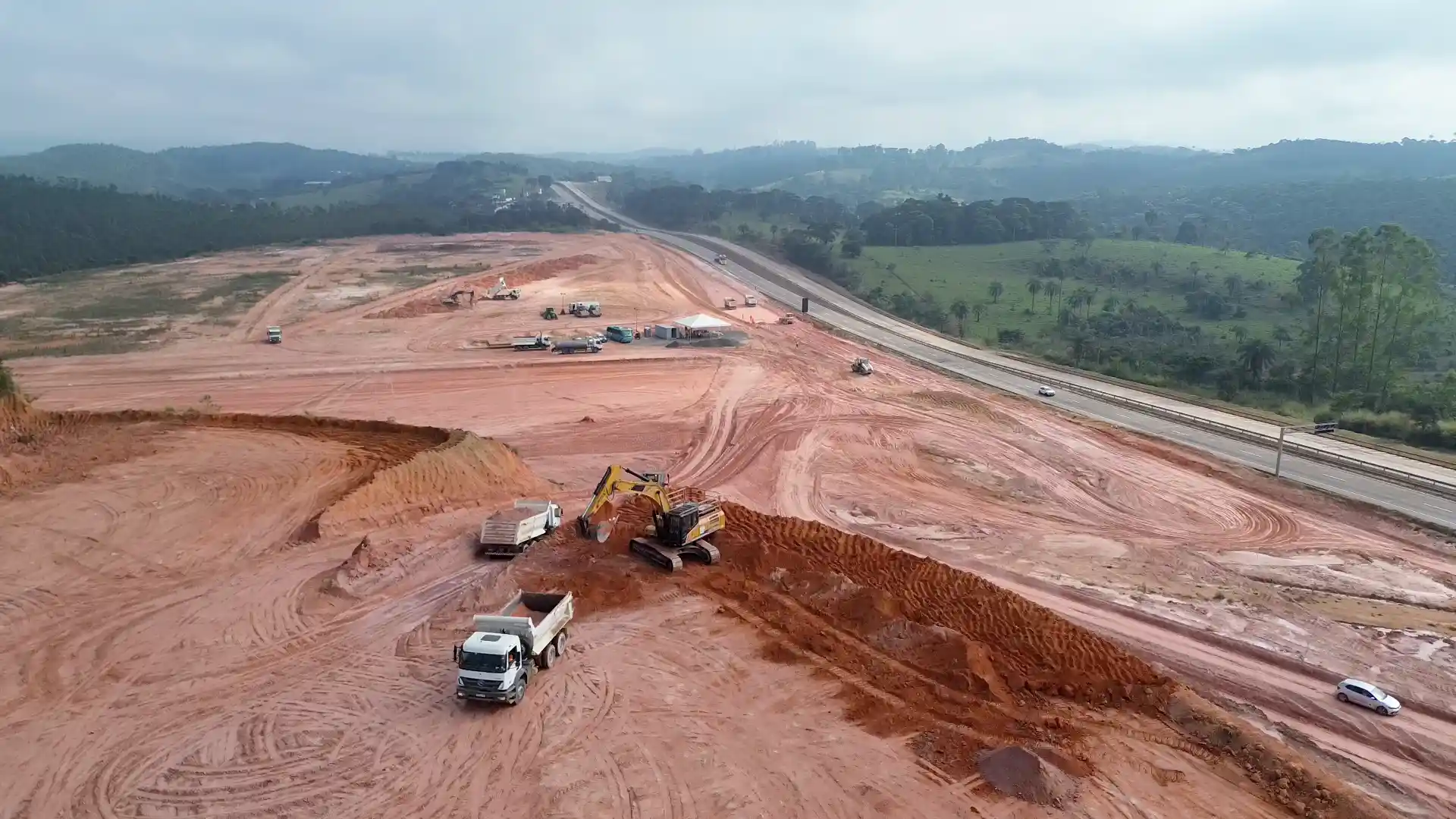 Vista aérea das obras do futuro Ponto de Parada e Descanso para caminhoneiros em Bom Jesus do Amparo, na BR-381, em Minas Gerais. Máquinas e caminhões realizam serviços de terraplanagem em uma ampla área de terra vermelha ao lado da rodovia, cercada por vegetação e colinas ao fundo.