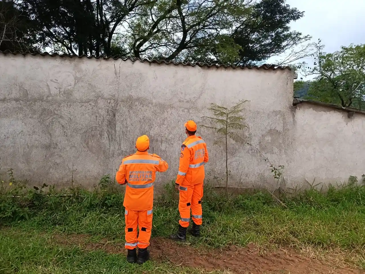 Imagem mostra dois bombeiros militares uniformizados observando uma área próxima a um muro no bairro Feixos, em João Monlevade. A equipe atuou na avaliação e eliminação de um enxame de abelhas que representava risco para participantes, organizadores e público de um evento esportivo realizado na região.