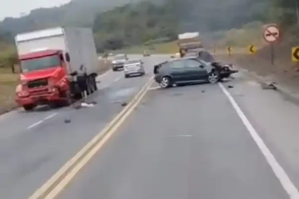 Imagem mostra acidente na BR-381. Um carro escuro está atravessado na pista e um caminhão vermelho com avarias na dianteira permanece parado no acostamento. Há destroços espalhados pela rodovia e algumas pessoas próximas aos veículos. O acidente ocorreu em um trecho cercado por vegetação e curvas.
