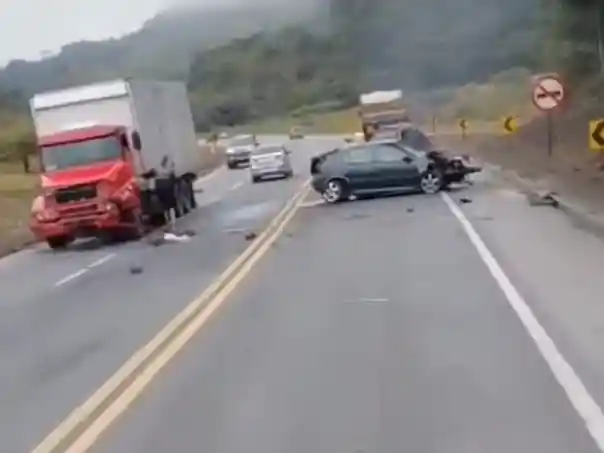 Imagem mostra acidente na BR-381. Um carro escuro está atravessado na pista e um caminhão vermelho com avarias na dianteira permanece parado no acostamento. Há destroços espalhados pela rodovia e algumas pessoas próximas aos veículos. O acidente ocorreu em um trecho cercado por vegetação e curvas.