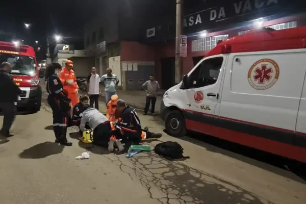 Socorristas do Sevor, bombeiros militares e profissionais do SAMU atendem uma vítima caída ao solo em uma via urbana de João Monlevade durante a noite. Ao lado da equipe estão estacionadas uma ambulância do SAMU e uma viatura do Corpo de Bombeiros, enquanto moradores acompanham a ocorrência à distância.
