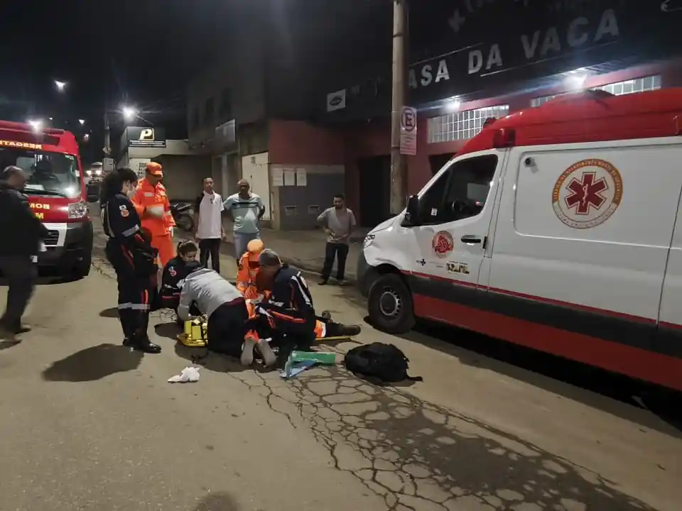 Socorristas do Sevor, bombeiros militares e profissionais do SAMU atendem uma vítima caída ao solo em uma via urbana de João Monlevade durante a noite. Ao lado da equipe estão estacionadas uma ambulância do SAMU e uma viatura do Corpo de Bombeiros, enquanto moradores acompanham a ocorrência à distância.