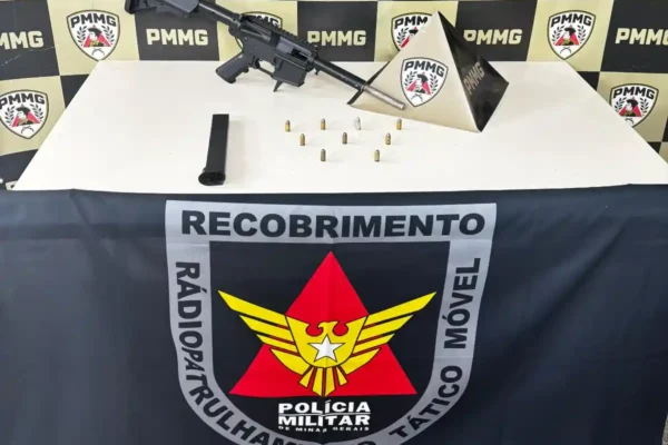 Imagem mostra uma submetralhadora artesanal, um carregador e diversas munições apreendidas pela Polícia Militar em uma residência no bairro Cruzeiro Celeste, em João Monlevade. Os materiais estão sobre uma mesa, em frente a um painel com símbolos da PMMG e da equipe de Radiopatrulhamento Tático Móvel.