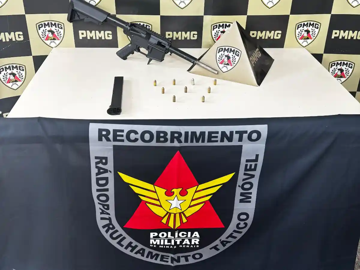 Imagem mostra uma submetralhadora artesanal, um carregador e diversas munições apreendidas pela Polícia Militar em uma residência no bairro Cruzeiro Celeste, em João Monlevade. Os materiais estão sobre uma mesa, em frente a um painel com símbolos da PMMG e da equipe de Radiopatrulhamento Tático Móvel.