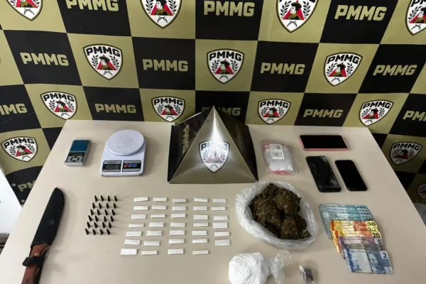 Itens apreendidos em uma ação da Polícia Militar estão dispostos sobre uma superfície, incluindo porções de cocaína e maconha, dinheiro, balanças de precisão, celulares e materiais para embalagem de entorpecentes. A ocorrência foi registrada no bairro Lucília, em João Monlevade, e resultou na prisão de um jovem suspeito de tráfico de drogas.