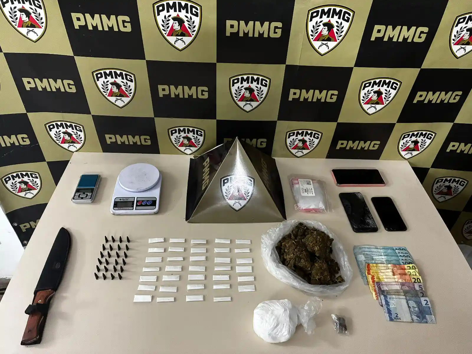 Itens apreendidos em uma ação da Polícia Militar estão dispostos sobre uma superfície, incluindo porções de cocaína e maconha, dinheiro, balanças de precisão, celulares e materiais para embalagem de entorpecentes. A ocorrência foi registrada no bairro Lucília, em João Monlevade, e resultou na prisão de um jovem suspeito de tráfico de drogas.
