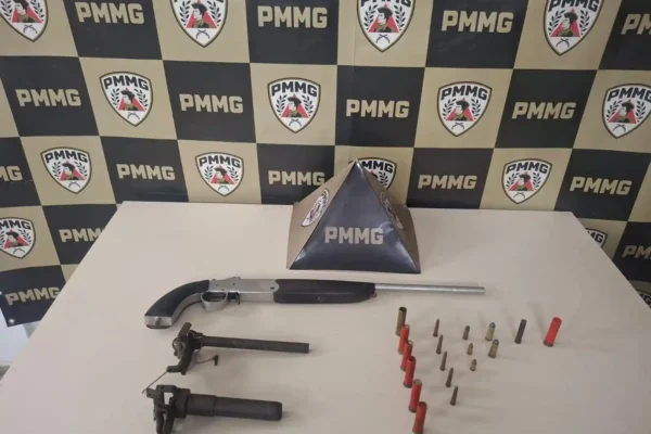 Imagem mostra materiais apreendidos pela Polícia Militar no bairro Laranjeiras, em João Monlevade. Sobre uma mesa branca estão três armas de fogo e várias munições organizadas em fileiras. Ao fundo, há um painel com os símbolos e a identificação da PMMG, utilizado para registros de ocorrências policiais.