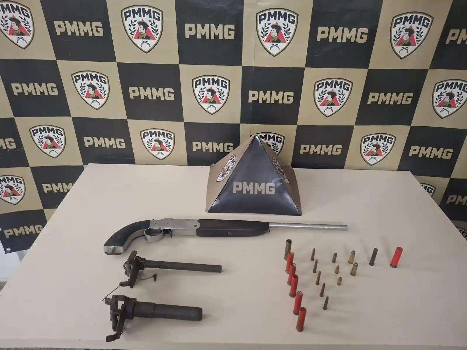 Imagem mostra materiais apreendidos pela Polícia Militar no bairro Laranjeiras, em João Monlevade. Sobre uma mesa branca estão três armas de fogo e várias munições organizadas em fileiras. Ao fundo, há um painel com os símbolos e a identificação da PMMG, utilizado para registros de ocorrências policiais.