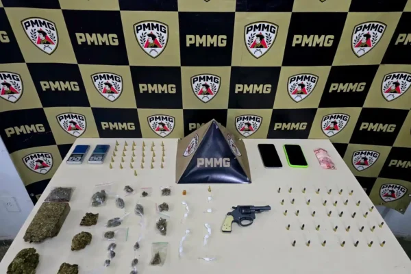 Materiais apreendidos pela Polícia Militar, no bairro Novo Cruzeiro, estão organizados sobre uma mesa diante de um painel da corporação. Entre os itens estão um revólver calibre .22, munições, porções de maconha, cocaína e crack, balanças de precisão, aparelhos celulares e dinheiro em espécie.