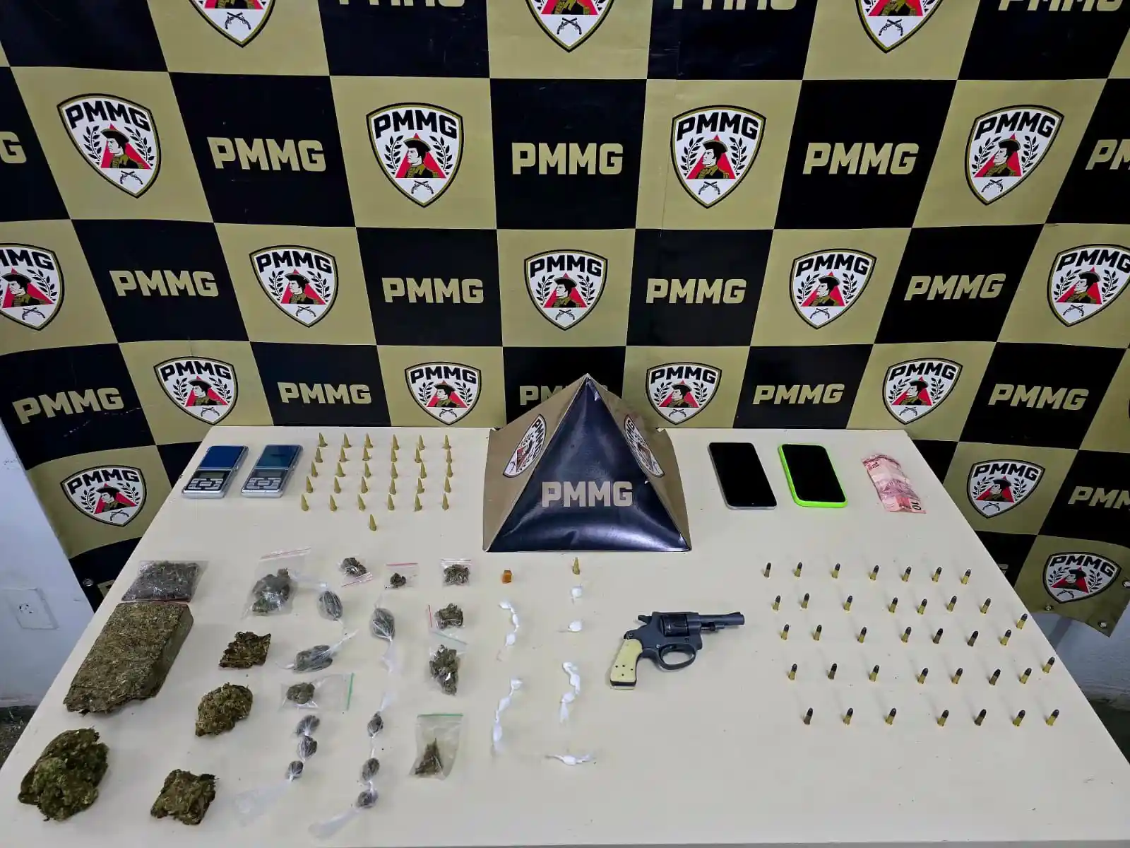 Materiais apreendidos pela Polícia Militar, no bairro Novo Cruzeiro, estão organizados sobre uma mesa diante de um painel da corporação. Entre os itens estão um revólver calibre .22, munições, porções de maconha, cocaína e crack, balanças de precisão, aparelhos celulares e dinheiro em espécie.
