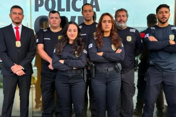 Sete integrantes da Polícia Civil estão posicionados em frente à fachada da delegacia. Os policiais vestem uniformes pretos, enquanto um deles usa terno escuro com gravata vermelha e distintivo. Ao fundo, aparece a identificação da Polícia Civil no prédio.