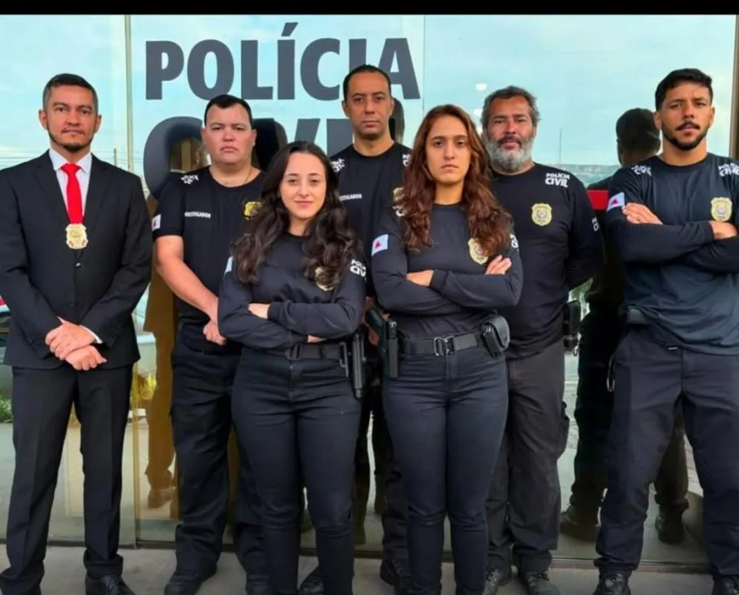 Sete integrantes da Polícia Civil estão posicionados em frente à fachada da delegacia. Os policiais vestem uniformes pretos, enquanto um deles usa terno escuro com gravata vermelha e distintivo. Ao fundo, aparece a identificação da Polícia Civil no prédio.