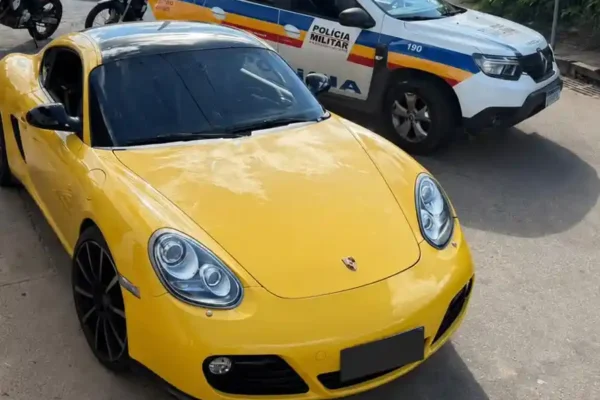 Fotografia mostra um Porsche Cayman amarelo estacionado em via pública ao lado de uma viatura da Polícia Militar de Minas Gerais. O veículo foi apreendido durante ocorrência registrada no bairro Cruzeiro Celeste, em João Monlevade, envolvendo embriaguez ao volante, direção perigosa e desobediência à ordem de parada policial.