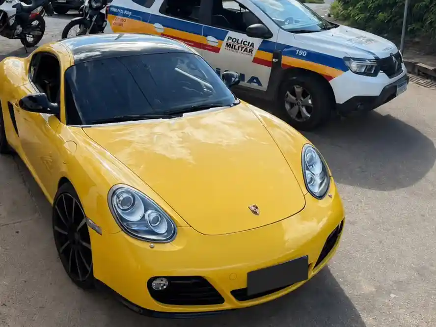 Fotografia mostra um Porsche Cayman amarelo estacionado em via pública ao lado de uma viatura da Polícia Militar de Minas Gerais. O veículo foi apreendido durante ocorrência registrada no bairro Cruzeiro Celeste, em João Monlevade, envolvendo embriaguez ao volante, direção perigosa e desobediência à ordem de parada policial.