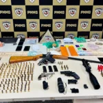 Mesa com armas, munições, drogas, dinheiro, celulares e equipamentos eletrônicos apreendidos pela Polícia Militar durante a Operação Luminis. Ao fundo, painel com a identidade visual da PMMG destaca a apresentação dos materiais recolhidos em ações realizadas em Nova Era, João Monlevade e São José do Goiabal.