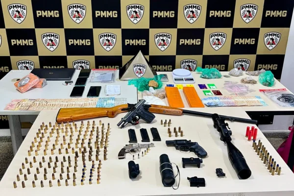 Mesa com armas, munições, drogas, dinheiro, celulares e equipamentos eletrônicos apreendidos pela Polícia Militar durante a Operação Luminis. Ao fundo, painel com a identidade visual da PMMG destaca a apresentação dos materiais recolhidos em ações realizadas em Nova Era, João Monlevade e São José do Goiabal.