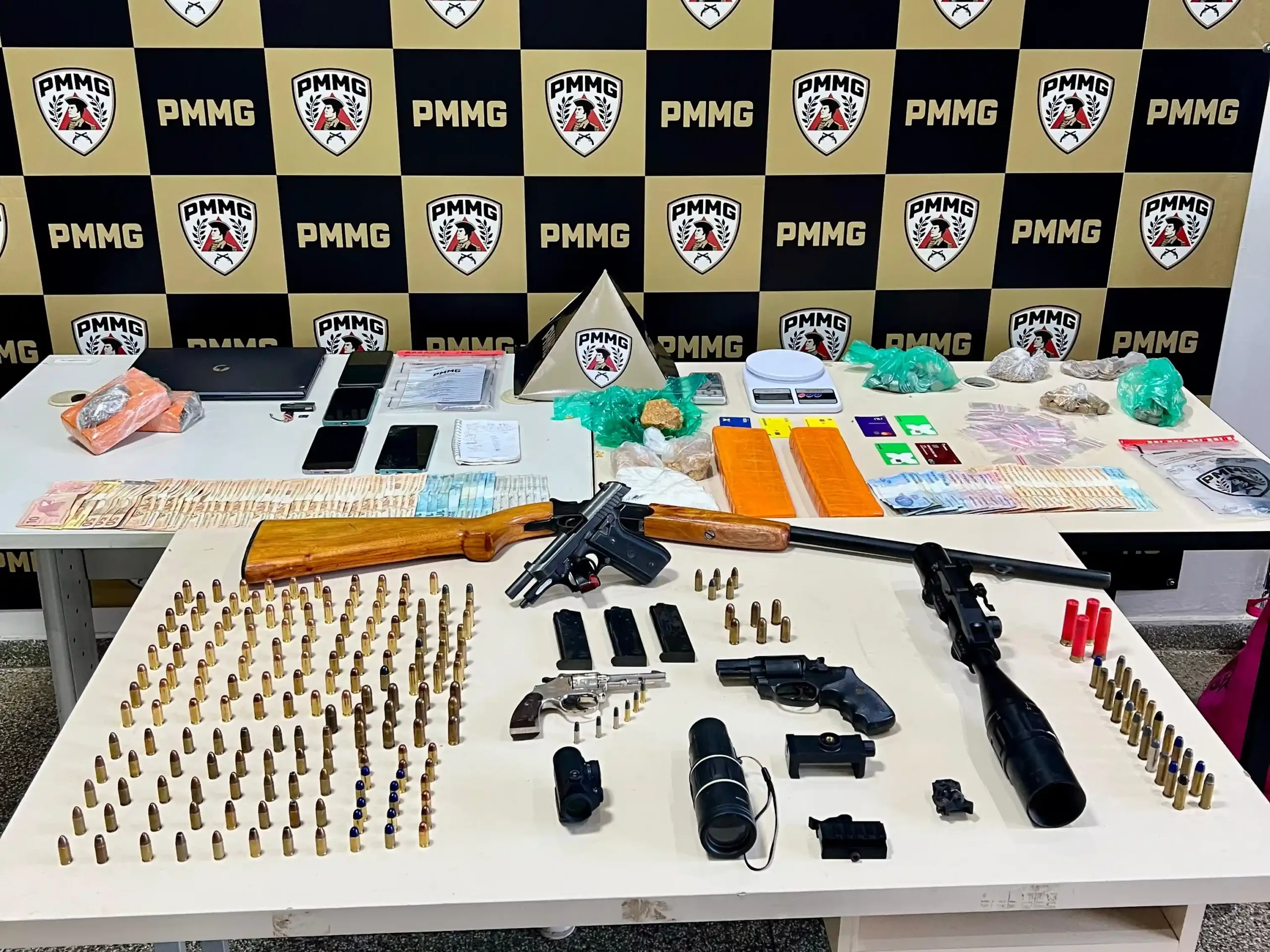 Mesa com armas, munições, drogas, dinheiro, celulares e equipamentos eletrônicos apreendidos pela Polícia Militar durante a Operação Luminis. Ao fundo, painel com a identidade visual da PMMG destaca a apresentação dos materiais recolhidos em ações realizadas em Nova Era, João Monlevade e São José do Goiabal.