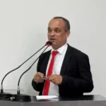 O vereador Bruno Cabeção aparece em pé, atrás de uma tribuna, durante pronunciamento em sessão legislativa. Vestindo terno escuro, camisa branca e gravata vermelha.