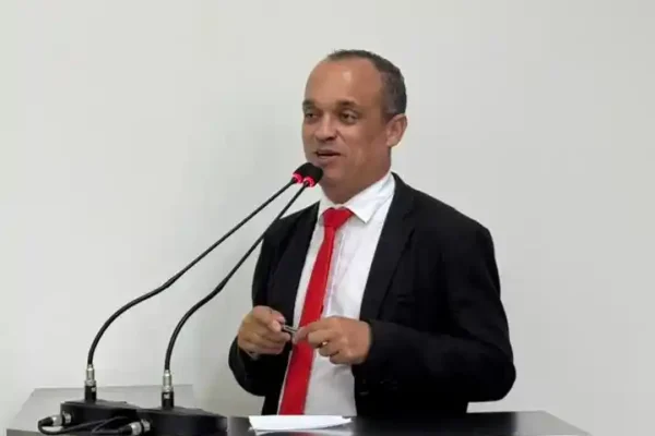 O vereador Bruno Cabeção aparece em pé, atrás de uma tribuna, durante pronunciamento em sessão legislativa. Vestindo terno escuro, camisa branca e gravata vermelha.