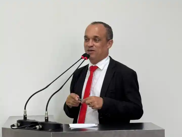 O vereador Bruno Cabeção aparece em pé, atrás de uma tribuna, durante pronunciamento em sessão legislativa. Vestindo terno escuro, camisa branca e gravata vermelha.