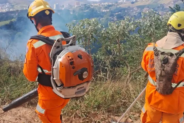 Dois profissionais do Corpo de Bombeiros e Brigada Florestal utilizam equipamentos de combate a incêndios em uma área de vegetação atingida pelo fogo em João Monlevade. Ao fundo, é possível observar fumaça saindo da área afetada e parte da paisagem urbana da cidade.