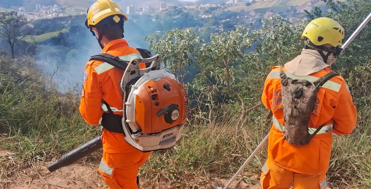 Dois profissionais do Corpo de Bombeiros e Brigada Florestal utilizam equipamentos de combate a incêndios em uma área de vegetação atingida pelo fogo em João Monlevade. Ao fundo, é possível observar fumaça saindo da área afetada e parte da paisagem urbana da cidade.