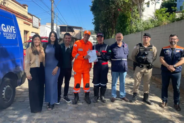 Integrantes do GAVE, Corpo de Bombeiros, Polícia Militar, Defesa Civil e representantes do poder público posam para foto durante cerimônia de entrega do certificado de credenciamento do GAVE, em Nova Era. Ao fundo, aparece uma viatura da instituição e, ao centro, participantes seguram o documento oficial concedido pelo Corpo de Bombeiros Militar de Minas Gerais.