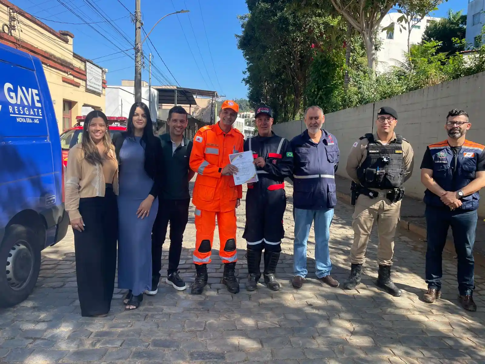 Integrantes do GAVE, Corpo de Bombeiros, Polícia Militar, Defesa Civil e representantes do poder público posam para foto durante cerimônia de entrega do certificado de credenciamento do GAVE, em Nova Era. Ao fundo, aparece uma viatura da instituição e, ao centro, participantes seguram o documento oficial concedido pelo Corpo de Bombeiros Militar de Minas Gerais.