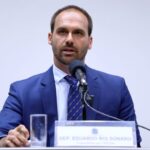 O ex-deputado federal Eduardo Bolsonaro aparece sentado atrás de uma mesa, usando terno azul e gravata. Ele fala ao microfone durante uma audiência ou reunião.