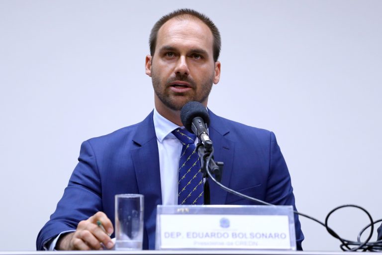 O ex-deputado federal Eduardo Bolsonaro aparece sentado atrás de uma mesa, usando terno azul e gravata. Ele fala ao microfone durante uma audiência ou reunião.
